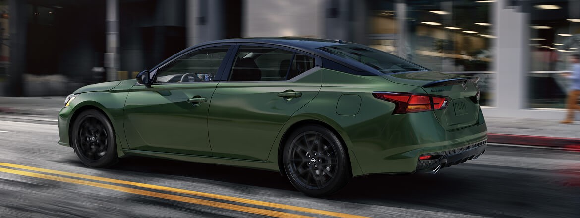 2026 Nissan Altima 3/4 rear side view of Altima driving on a city road with modern building in background.